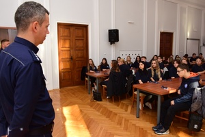 Policjanci podczas spotkania z uczniami. Policjanci stoją, uczniowie siedzą przy długich stołach.