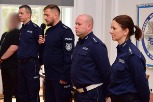 Policjanci podczas spotkania z uczniami. Policjanci stoją, uczniowie siedzą przy długich stołach.