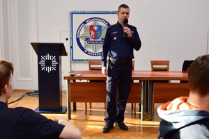 Policjanci podczas spotkania z uczniami. Policjanci stoją, uczniowie siedzą przy długich stołach.