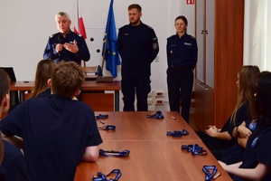 Policjanci podczas spotkania z uczniami. Policjanci stoją, uczniowie siedzą przy długich stołach.