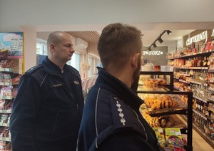 Policjanci kontrolujący sprzedaż alkoholu dzieciom w sklepie, podczas rozmowy z personelem placówki. W tle wyposażenie sklepu, wyłożony na pólkach towar