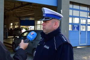 Policjant udziela wywiadu dziennikarzowi przed budynkiem Okręgowej Stacji Kontroli Pojazdów. W tle za funkcjonariuszem widać pojazdy przechodzące badania techniczne