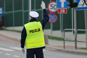 policjant stojący tyłem do obiektywu w odblaskowej kamizelce, z uniesioną prawa ręką do góry, w czasie regulacji ruchem na drodze