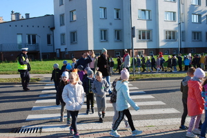 Dzieci przechodzą przez przejście dla pieszych. Obok funkcjonariusz ruchu drogowego kieruje ruchem.