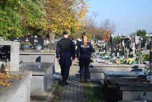 Na zdjęciu tyłem policjantka i policjant umundurowani w granatowe mundury patrolują teren cmentarza. Policjanci idą alejka pomiędzy nagrobkami.