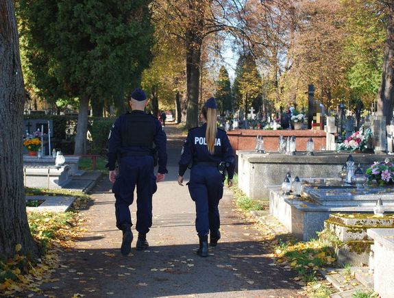 Na zdjęciu tyłem policjantka i policjant umundurowani w granatowe mundury patrolują teren cmentarza. Policjanci idą alejka pomiędzy nagrobkami.