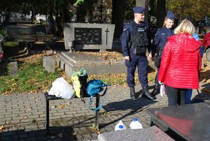 Umundurowani policjanci podczas patrolu na terenie cmentarza rozmawiają z kobietami porządkującymi nagrobek o bezpieczeństwie. Funkcjonariusze zwracają uwagę na pozostawione bez nadzoru torebki.