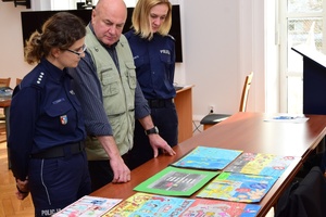 Policjanci i zaproszeni członkowie jury przeglądają prace konkursowe rozłożone na stołach.