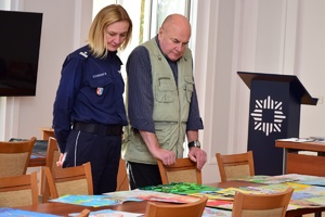 Policjanci i zaproszeni członkowie jury przeglądają prace konkursowe rozłożone na stołach.