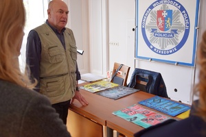 Policjanci i zaproszeni członkowie jury przeglądają prace konkursowe rozłożone na stołach.