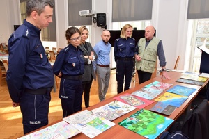 Policjanci i zaproszeni członkowie jury przeglądają prace konkursowe rozłożone na stołach.