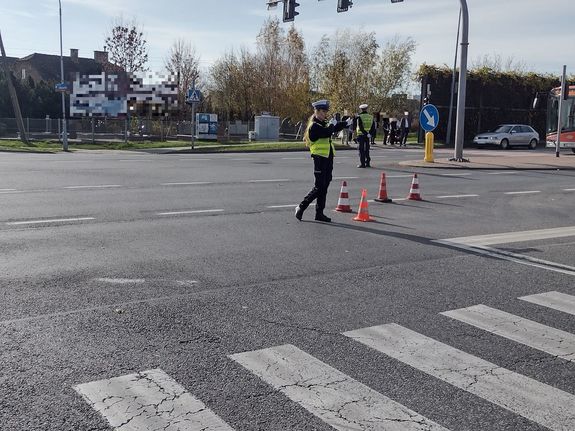 Na zdjęciu stojąca przodem umundurowana policjantka ruchu drogowego kierująca ruchem na skrzyżowaniu ulicy Lwowskiej z Cienistą. W dalszym planie stojący plecami policjant ruchu drogowego. Jadący ulicą Lwowską samochód osobowy oraz autobus. Przed przejściem dla pieszych stoją piesi.
