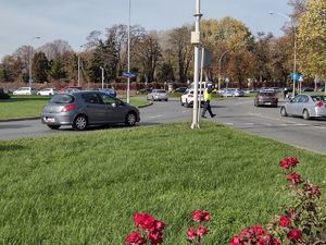 Na zdjęciu stojący bokiem umundurowany policjant ruchu drogowego kierujący ruchem na rondzie Pobito, na pasach ruchu przejeżdżające samochody. W dalszym planie przechodzący na pasach piesi i cmentarz.