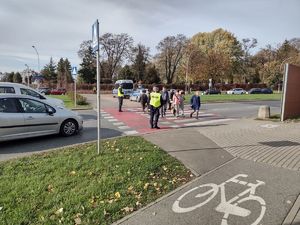 Na zdjęciu przejście dla pieszych, dwóch umundurowanych funkcjonariuszy: jeden ruchu drogowego, drugi prewencji oraz piesi. Przed przejściem jadące ulica pojazdy. W tle cmentarz.