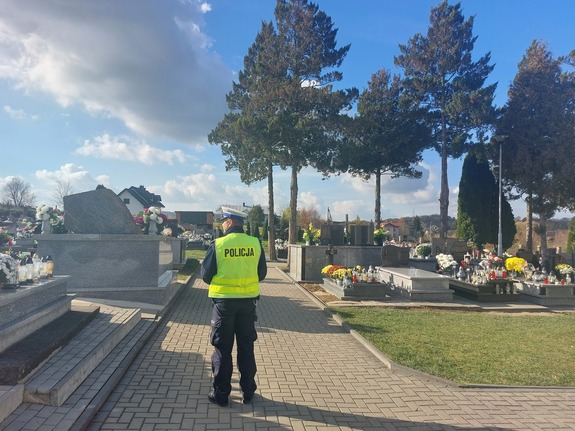 Na zdjęciu widoczny umundurowany policjant stojący w rejonie cmentarza w związku z akcją &quot;Znicz&quot;, dbanie o bezpieczeństwo w ruchu drogowym i kontrola prędkości.
