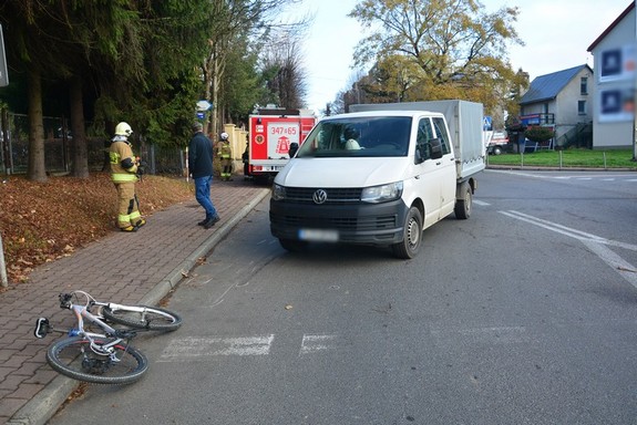 Leżący na jezdni rower, tuż obok niego biały dostawczy volkswagen. W tle strażak, świadek zdarzenia i wóz strażacki