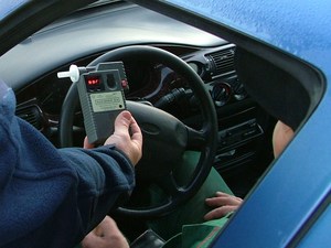 Policjant trzymający w ręku urządzenie do badania stanu trzeźwości. Wynik badania pokazuje kierowcy, który siedzi w aucie i ma odsunięta przednia szybę