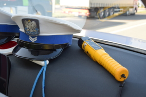 Policyjna czapka i urządzenie do sprawdzania stanu trzeźwości położone na desce rozdzielczej radiowozu. Przez szybę radiowozu widać samochód ciężarowy