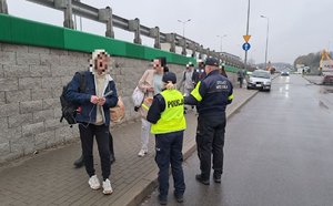 Na zdjeciu widać wiadukt w ciągu drogi krajowej nr 9 i Poniżej wiaduktu na chodniku umundurowana policjantka i funkcjonariusz strazy miejskiej rozdają odblaskowe opaski grupie pieszych idących chodnikiem.