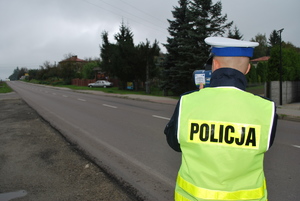 zdjęcie przedstawia umundurowanego policjanta w trakcie mierzenia prędkości pojazdu poruszającego się po drodze