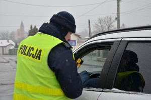 Policjant stojący tyłem do obiektywu w seledynowej kamizelce odblaskowej z napisem Policja, trzymając w ręku urządzenie do badania stanu trzeźwości podczas kontroli drogowej przy srebrnym samochodzie