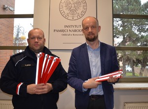 Po lewej policjant z biało-czerwonymi opaskami odblaskowymi w rękach. Po prawej przedstawiciel IPN trzymający odblaski. W tle logo IPN.