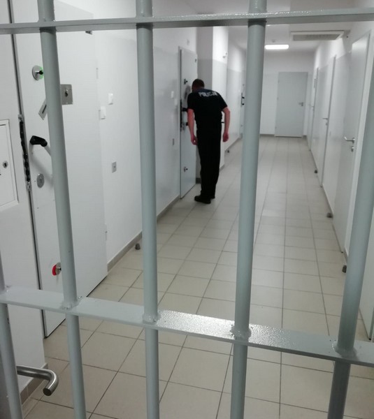 Krata w kolorze szarym, za którą znajduje się korytarz pomieszczeń dla osób zatrzymanych. W tle policjant dozorujący przez wizjer osobę zatrzymaną