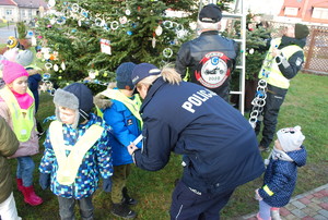 Na zdjęciu policjantka ubrana w granatowy mundur z napisem policja na plecach zakłada dzieciom odblaski. Przed policjantkom stoi kilkoro przedszkolaków. Na drugim planie wolontariusze ozdabiają choinkę.