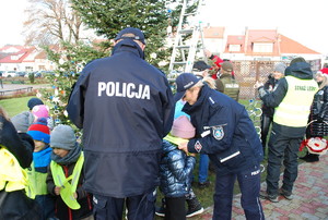 Na zdjęciu policjantka i policjant ubrani w granatowe mundury z napisem policja na plecach zakładają dzieciom odblaski. Przed policjantkom stoi kilkoro przedszkolaków. Na drugim planie wolontariusze ozdabiają choinkę.