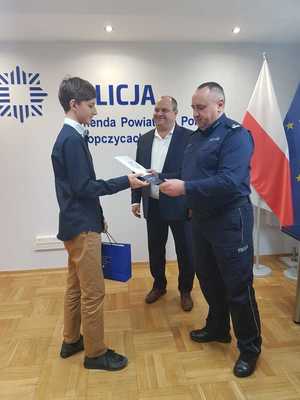 Po lewej stronie stoi laureat konkursu. Chłopiec odbiera nagrodę od podinsp. Romana Zawiślaka. Obok stoi Witold Darłak.