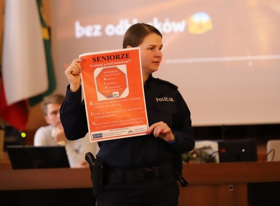Policjantka prezentująca plakat promujący kampanię „Seniorze – ucieknij przed oszustem”. Plakat w kolorze czerwonym z napisem w kolorze czarnym Seniorze, a poniżej białym – ucieknij przed oszustem