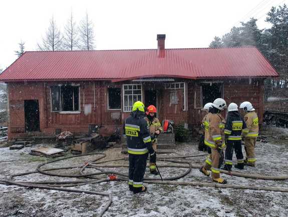 Spalony dom z otwartymi oknami przed nim strażacy zwijający węże pożarnicze.