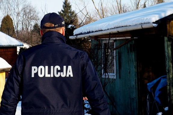 Policjant podczas patrolu ogródków działkowych w umundurowaniu służbowym z widocznym napisem policja na kurtce. Tło stanowi zimowa sceneria jednego z krośnieńskich ogródków
