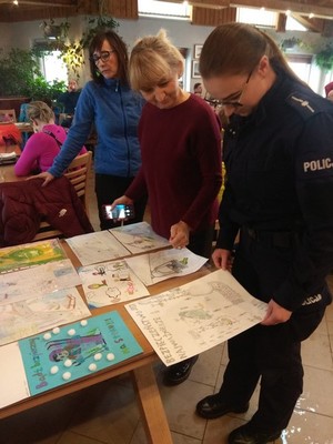 Policjantka oraz dwie członkinie komisji konkursowej, oceniają prace plastyczne o tematyce bezpieczeństwa w czasie zimowego wypoczynku. Kolorowe prace ułożone na drewnianych stołach