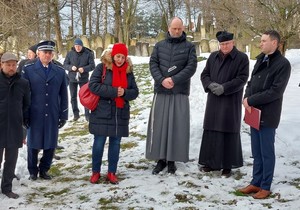 Osoby duchowne oraz uczestnicy modlitw na cmentarzy żydowskim w Krośnie. W tle cmentarne pomniki i drzewa. W lewej części fotografii, umundurowany policjant.