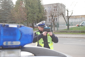 Policjant stojący przed radiowozem i mierzący prędkość jadących samochodów.