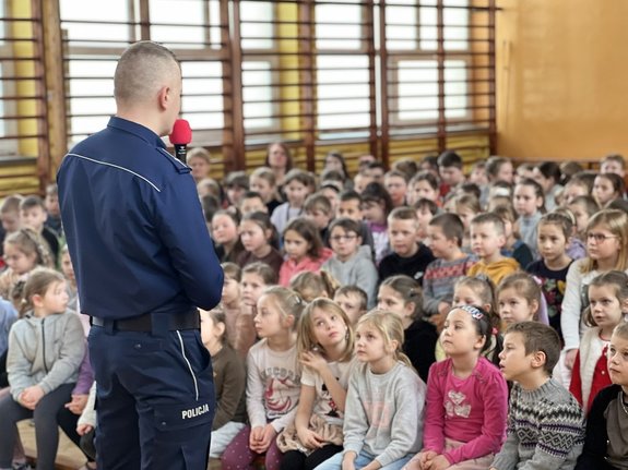 Zdjęcie przedstawia policjanta, który przy pomocy mikrofonu przeprowadza spotkanie z dziećmi na sali gimnastycznej.