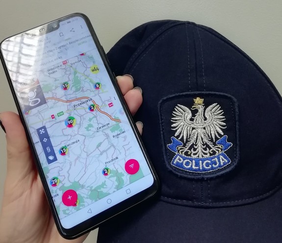 Telefon trzymany w ręce policjanta a na nim włączona aplikacji internetowa Krajowa Mapa Zagrożeń Bezpieczeństwa. Obok lezy na biurku czapka policyjna.