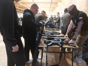 z lewej strony  zdjęcia widoczni  stojący chłopcy,  na środku widoczny stół na którym rozłożone są jednostki broni, z prawej strony stołu stoją policjanci w mundurach