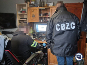 Dwóch policjantów, jeden siedzi, drugi stoi, w pokoju przed komputerem. Stojący ubrany w czarną kurtkę z białym napisem - CBZC