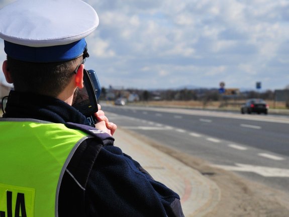 Na zdjęciu widoczny jest policjant wydziału ruchu drogowego stojący przy jezdni. Wykonuje on pomiar prędkości za pomocą ręcznego miernika prędkości.