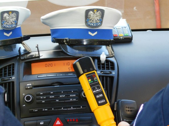 Na zdjęciu  widoczne urządzenie Alcoblow trzymane w ręce przez policjanta, na podszybiu radiowozu znajdują się dwie czapki służbowe z otokiem sierżanta policji, w tle terminal płatniczy,
