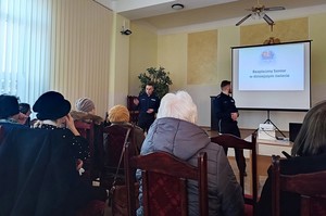 Funkcjonariusze podczas prelekcji w trakcie debaty w Wojaszówce.  W tle kadr z wyświetlanej prezentacji z tekstem „Bezpieczny senior w dzisiejszym świecie”
