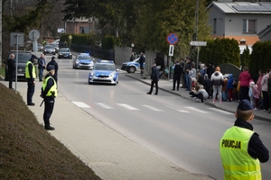 Policjanci podczas zabezpieczenia obchodów 79. rocznicy śmierci Rodziny Ulmów. Policjanci stoją wzdłuż ulicy, w centrum kadru jadący radiowóz a za nim kolumna pojazdów prezydenckich.