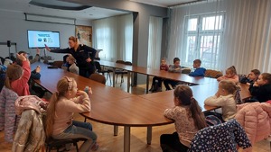 zdjęcia przedstawiają dzieci z klasy pierwszej na spotkaniu z policjantką w Komendzie Miejskiej Policji w Przemyślu. Uczniowie siedzą na auli przy stolikach i słuchają prelekcji policjantki.