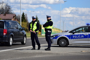 Dwóch policjantów stojących przy ulicy, przed radiowozem, a obok nich przejeżdża samochód. Pora dzienna