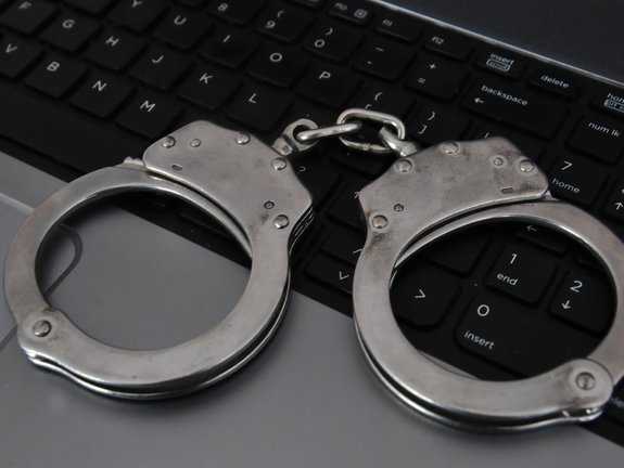 Na zdjęciu widoczne są policyjne kajdanki położone na klawiaturze komputera