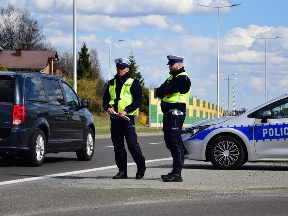 Na zdjęciu widoczni są dwaj funkcjonariusze ruchu drogowego stojący przy jezdni obok radiowozu