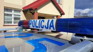 Baner z napisem policja na dachu radiowozu oznakowanego. W tle budynek koloru żółtego.