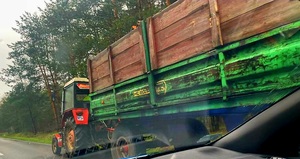 jezdnia przy lesie. na jezdni stoi ciągnik rolniczy koloru czerwonego  z przyczepą koloru zielono czerwonego